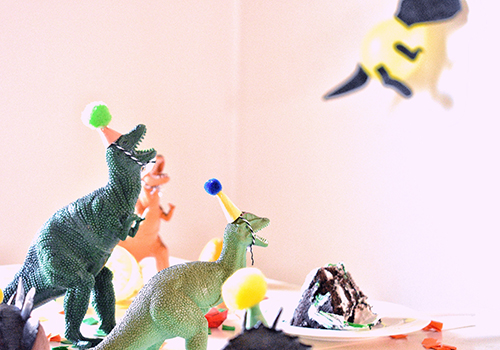  A shot of dinosaur figurines wearing party hats arranged around a slice of cake.
