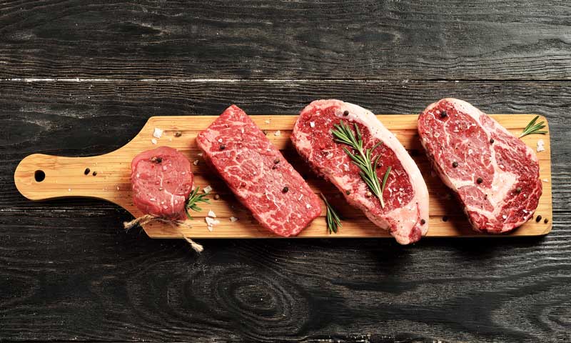 Different cuts of fresh beef laid on a wooden board with sprigs of rosemary and rock salt.
