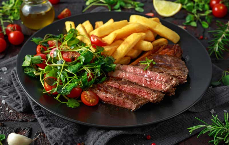 Medium rare steak with chips and salad on a stylish black plate.