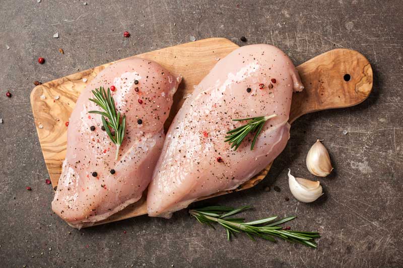 2 Raw chicken breasts on a wooden chopping board with sprigs of rosemary and cloves of garlic.