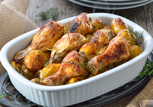 Roast chicken drumsticks in a casserole dish with roast potatoes, onions and sprigs of rosemary.