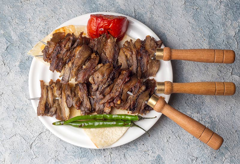 Lamb shish kebab on skewers placed on a plate with roasted peppers and chillies.