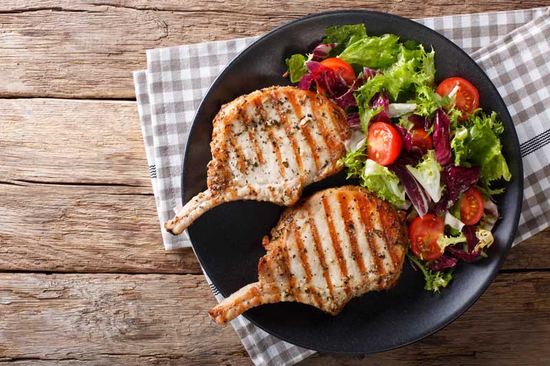 2  grilled pork chops with tomato and lettuce on a stylish black plate.