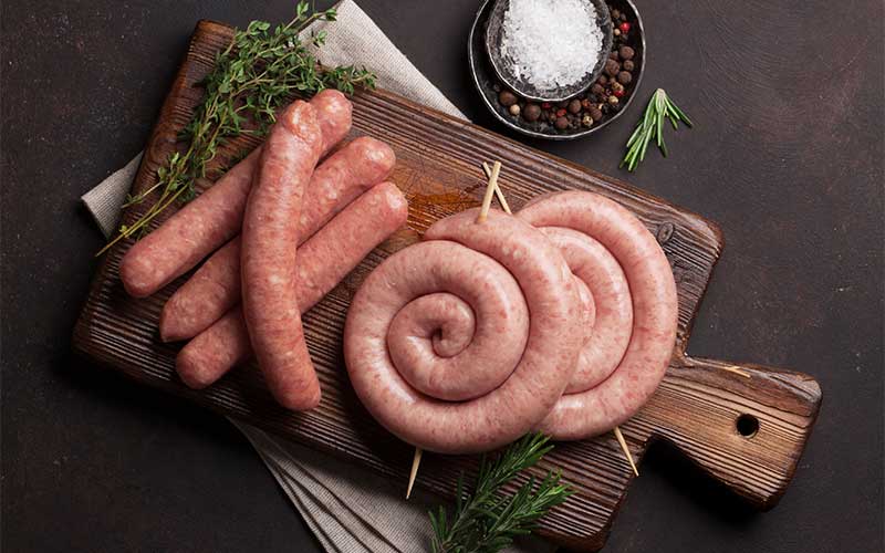 Raw sausages on a wooden chopping board with sprigs of rosemary and a dish of sea salt with peppercorns

