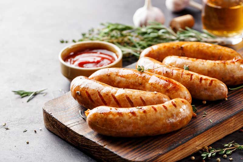 Grilled sausages resting on a wooden board with rosemary, garlic and a pot of tomato sauce.